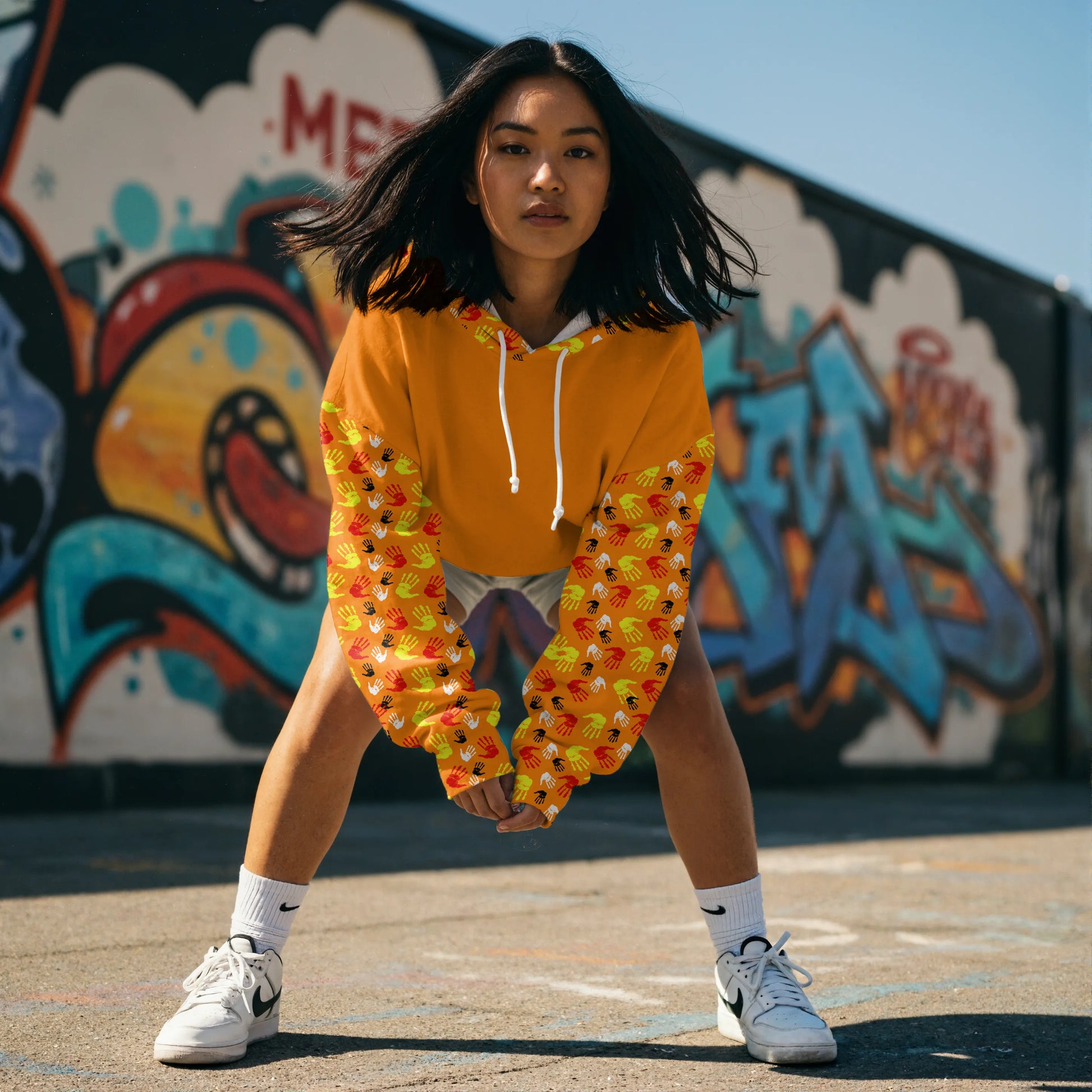 Indigenous woman outdoors wearing a bright orange pullover hoodie with a repeating handprint pattern on the sleeves, featuring a turtle graphic with colorful handprints on the back. The hoodie has the text "EVERY CHILD MATTERS" above the graphic, symbolizing awareness and unity for children’s rights.