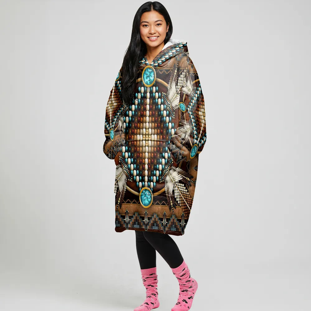 A woman standing front-facing in an oversized brown blanket hoodie decorated with turquoise geometric medallions, hands in pockets and smiling.