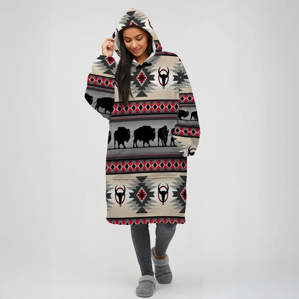 Woman modeling a beige hooded wearable blanket with horizontal stripes of black buffalo silhouettes, bull skull motifs, and red-and-gray southwestern diamond designs.