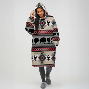 Woman modeling a beige hooded wearable blanket with horizontal stripes of black buffalo silhouettes, bull skull motifs, and red-and-gray southwestern diamond designs.