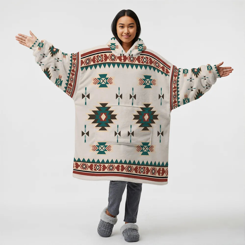 Person wearing an oversized cream hooded blanket covered in turquoise, red, and brown Native American–inspired geometric patterns, arms outstretched.