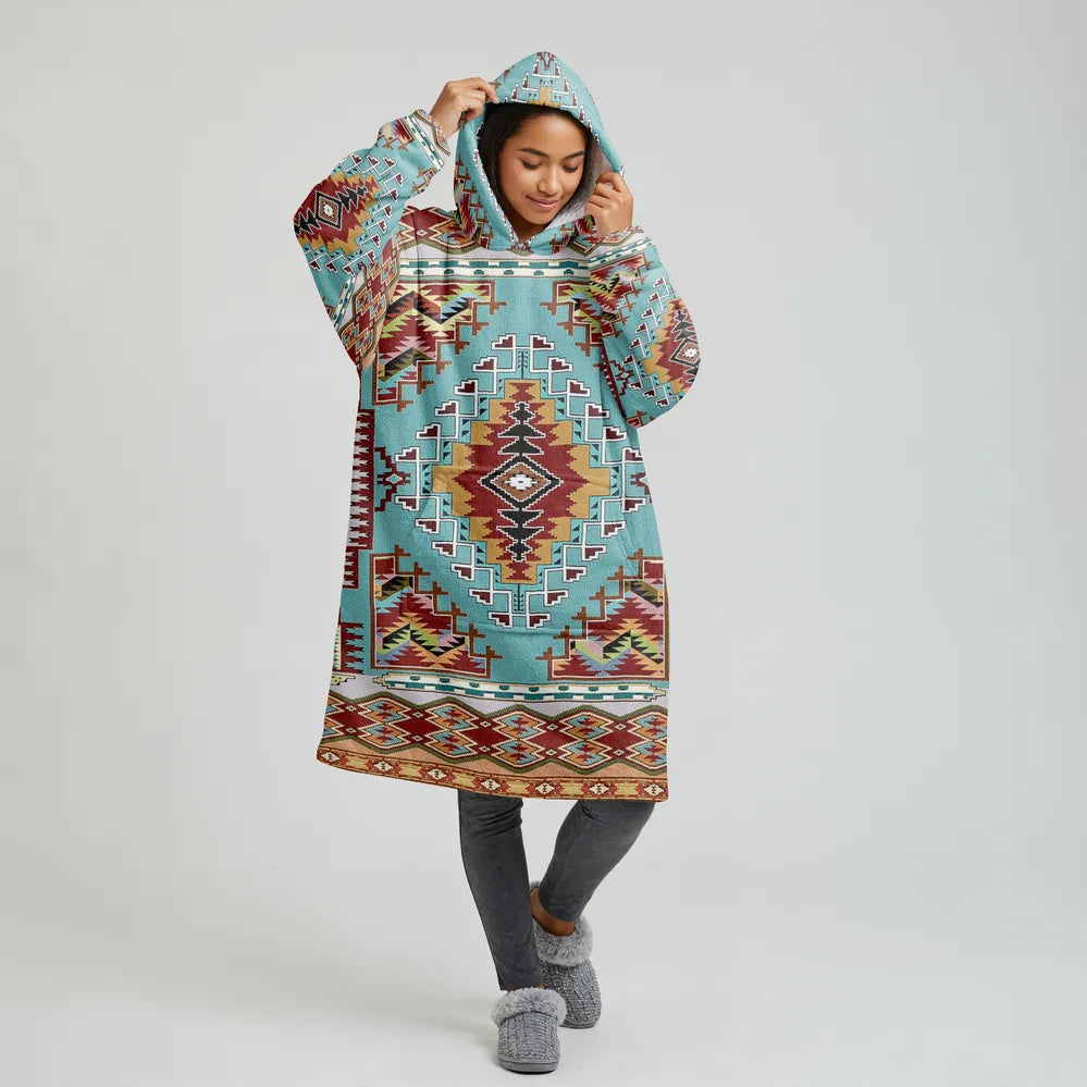 A young woman facing the camera wearing an oversized cream-colored wearable blanket hoodie patterned with teal and rust tribal motifs, hood up and adjusting it.