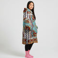 A woman wearing a cream and teal tribal-patterned blanket hoodie, with front pockets, black leggings, and pink socks. The design features vibrant geometric patterns.