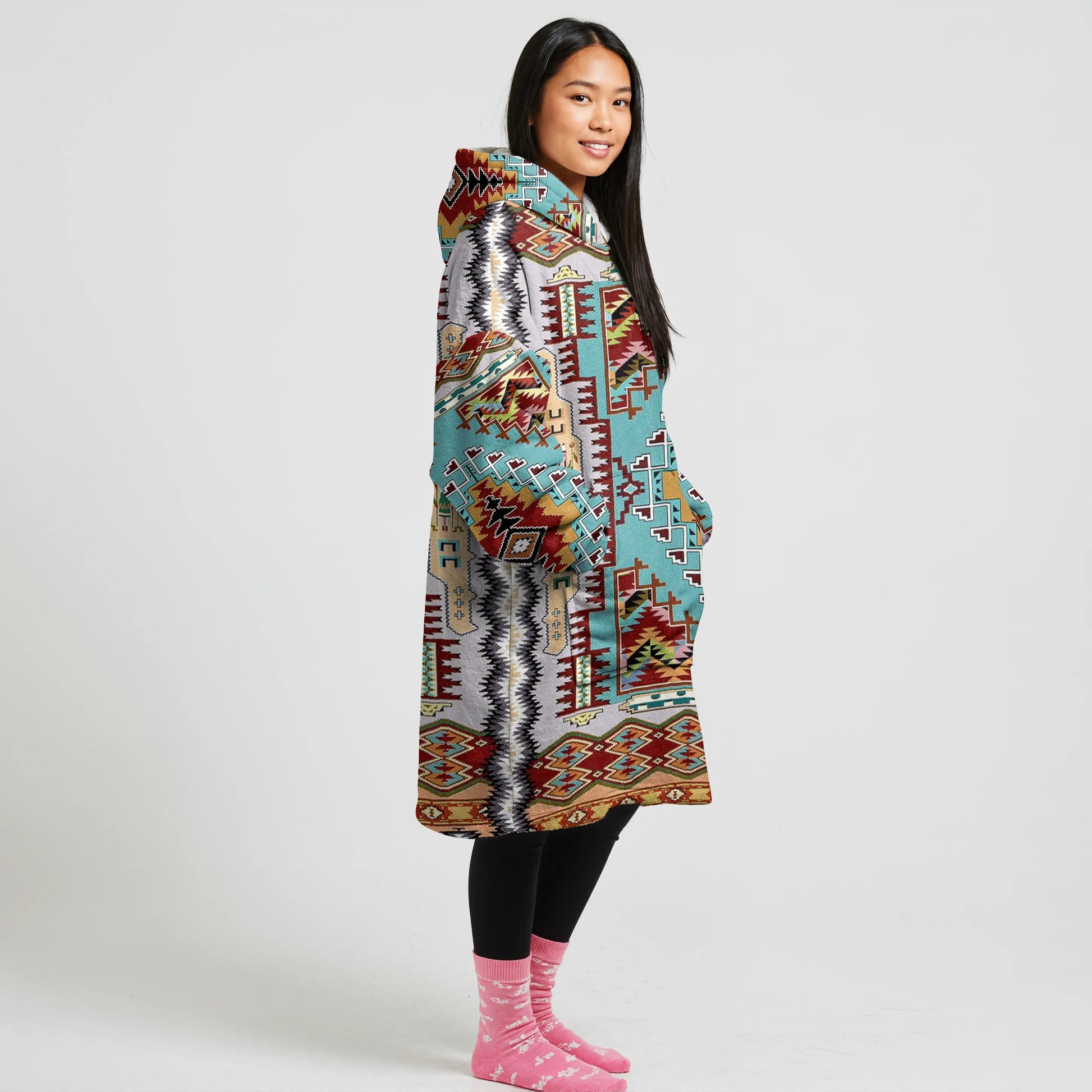 A woman wearing a cream and teal tribal-patterned blanket hoodie, with front pockets, black leggings, and pink socks. The design features vibrant geometric patterns.