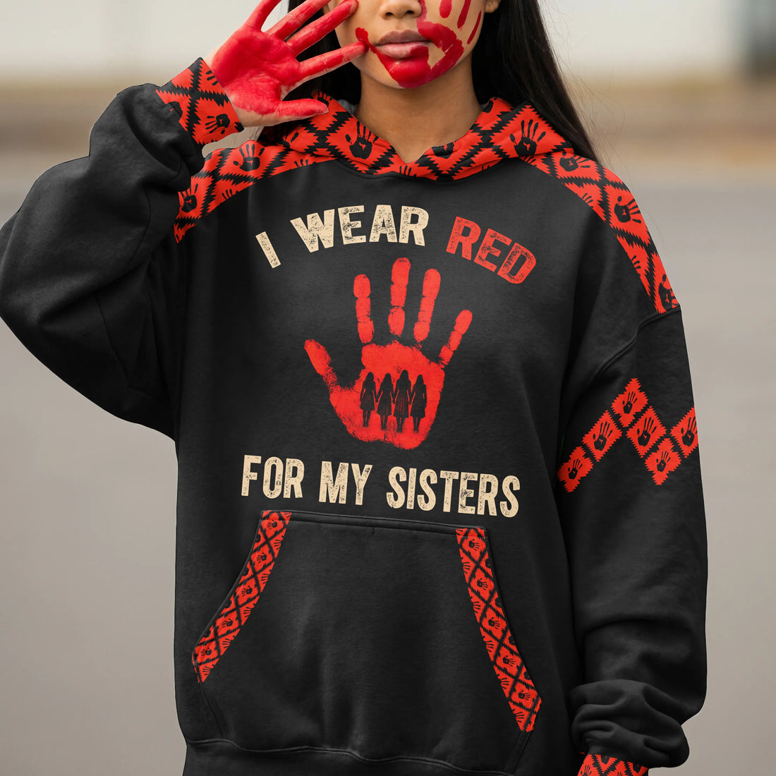 Close-up of a model’s hand and face painted red holding up the sleeve of a black hoodie, showcasing the red handprint graphic and “I Wear Red For My Sisters” text.