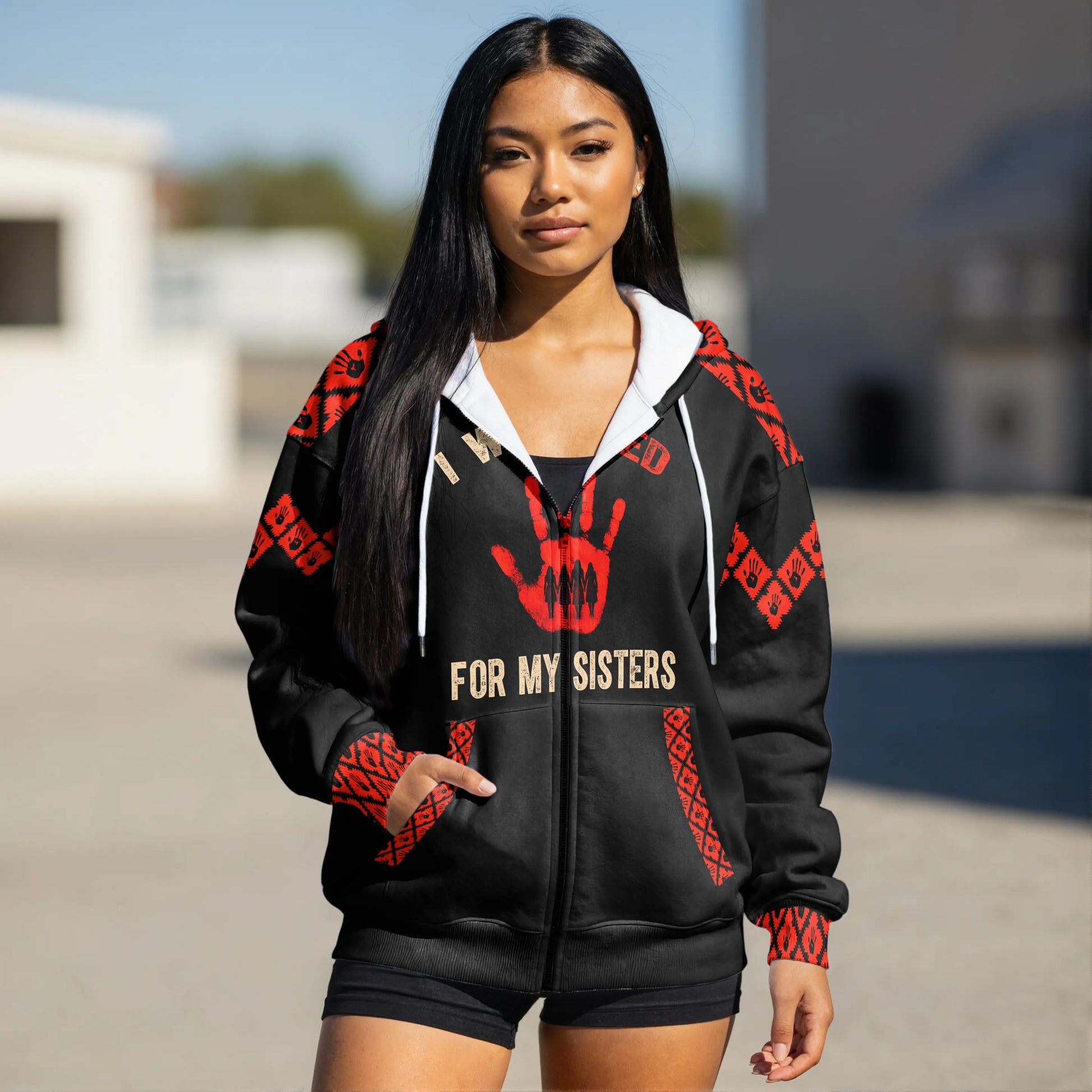 Front view of a woman taking a mirror selfie while wearing a black zip-up hoodie decorated with red tribal patterns on the shoulders and cuffs and a red handprint graphic reading “I Wear Red For My Sisters.