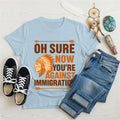 Light blue t‑shirt featuring brown chief headdress graphic and text “Oh Sure Now Youre Against Immigration.”
