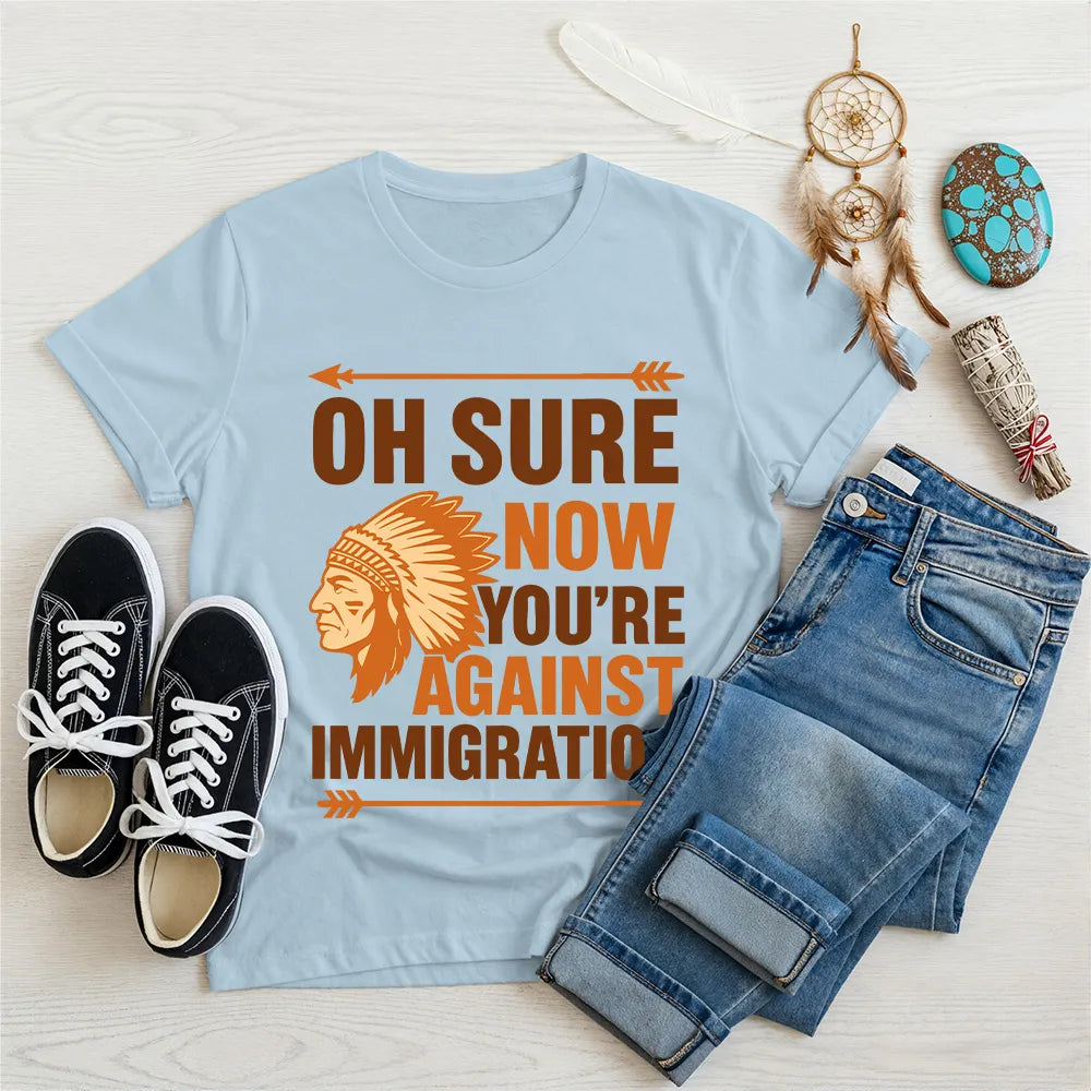 Light blue t‑shirt featuring brown chief headdress graphic and text “Oh Sure Now Youre Against Immigration.”