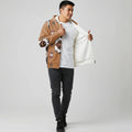 Male model in a tan Native-American–inspired zip-up jacket with graphic sleeve panels, shown open over a white tee, posing against a grey cyclorama.
