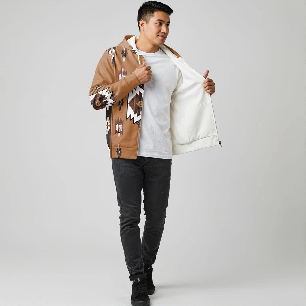 Male model in a tan Native-American–inspired zip-up jacket with graphic sleeve panels, shown open over a white tee, posing against a grey cyclorama.
