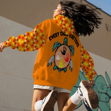 Indigenous woman jumping with her back to the camera, wearing an orange hoodie featuring a turtle design with colorful handprints on the back. The hoodie displays the words "Every Child Matters" above the turtle, and the sleeves are decorated with a pattern of colorful handprints. The background is a plain wall with minimal detail.