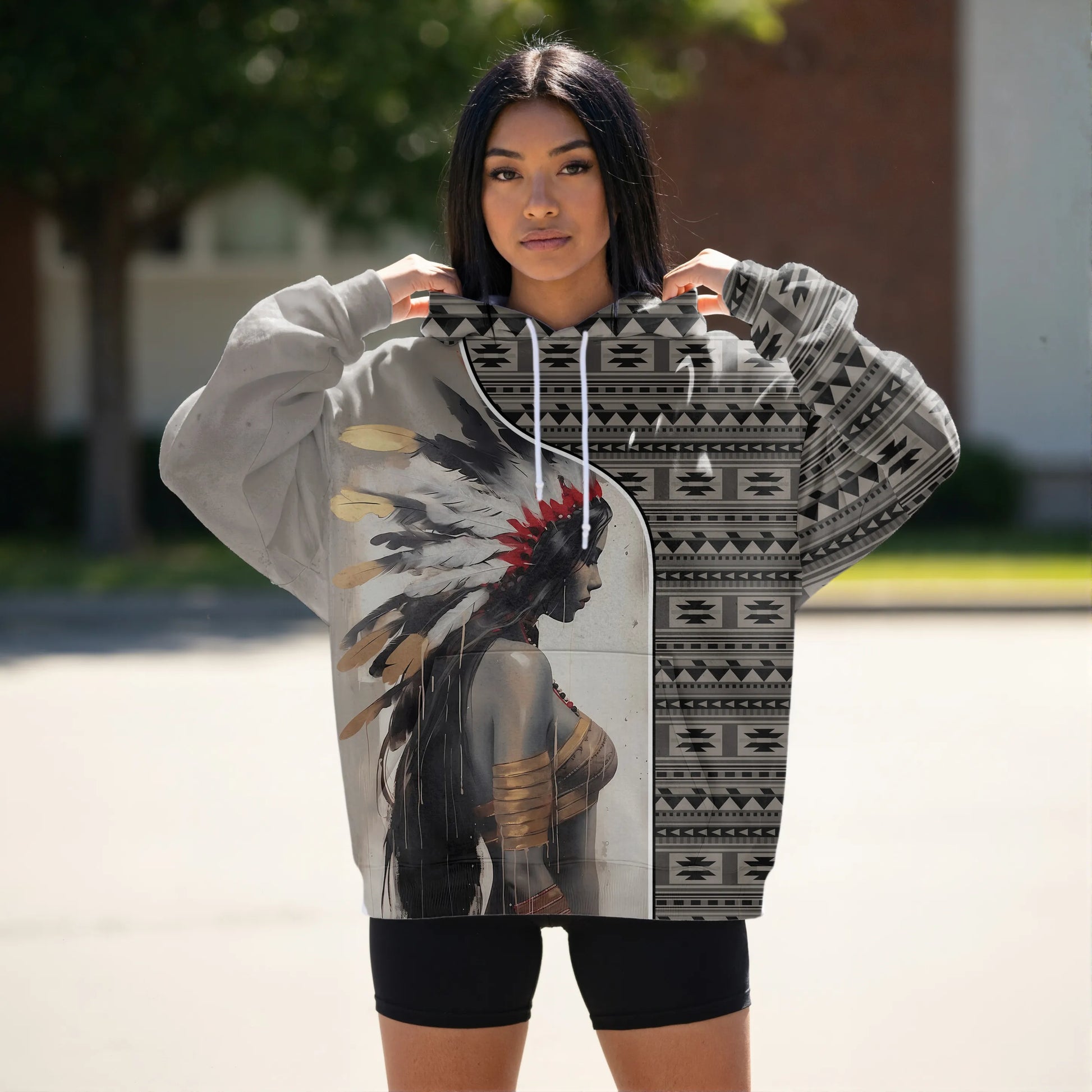 Young Indigenous woman outdoors wearing a bomber-style hoodie split vertically: on the left, a grayscale profile of a female warrior crowned with a feathered headdress; on the right, a Southwest tribal geometric pattern in monochrome tones, the hood and sleeves echoing the tribal motif.