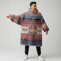 Model wearing an oversized earth-tone wearable blanket hoodie with authentic Native American–inspired tribal patterns and Sherpa lining, arms outstretched to showcase its roomy fit