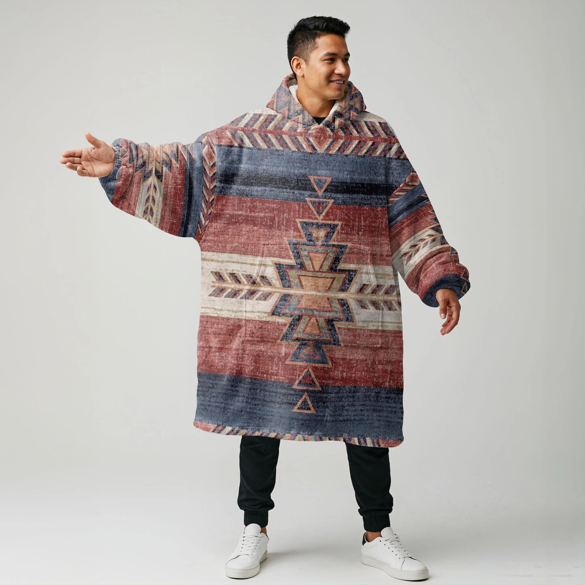 Model wearing an oversized earth-tone wearable blanket hoodie with authentic Native American–inspired tribal patterns and Sherpa lining, arms outstretched to showcase its roomy fit