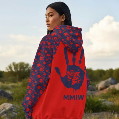 Indigenous woman standing outdoors in profile wearing a red and navy jacket with a repeated red handprint pattern on the sleeves and hood and a large navy handprint graphic featuring a female face silhouette and the letters “MMIW” on the back.