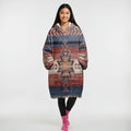 Seated model in a reversible wearable blanket jacket featuring southwestern tribal motifs in warm browns and reds, finished with plush Sherpa lining