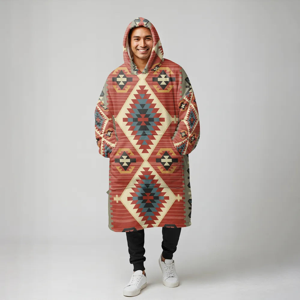 A man facing the camera wearing an oversized rust-colored blanket hoodie patterned with teal and cream diamond motifs, hands in the front pocket.