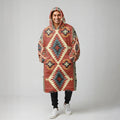 A man facing the camera wearing an oversized rust-colored blanket hoodie patterned with teal and cream diamond motifs, hands in the front pocket.