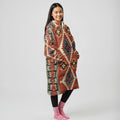Side profile of a woman in a rust-colored diamond-patterned blanket hoodie, her hand casually in the pocket, showing the garment’s drape and sleeve details.