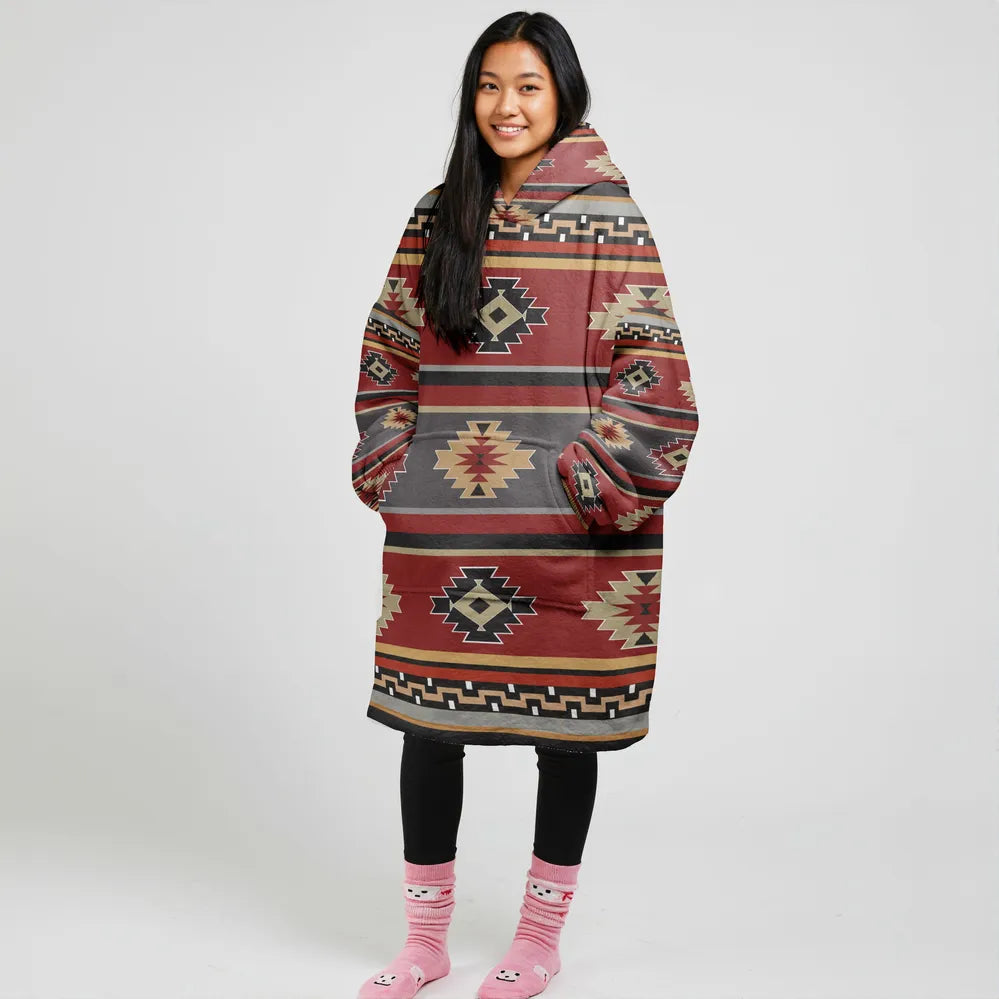 Woman standing in a red and brown wearable blanket hoodie with tribal diamond patterns and fringe details, showcasing its oversized, cozy design