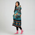 A woman front-facing in an oversized teal and coral striped tribal blanket hoodie, hands tucked into the pocket, smiling at the camera.
