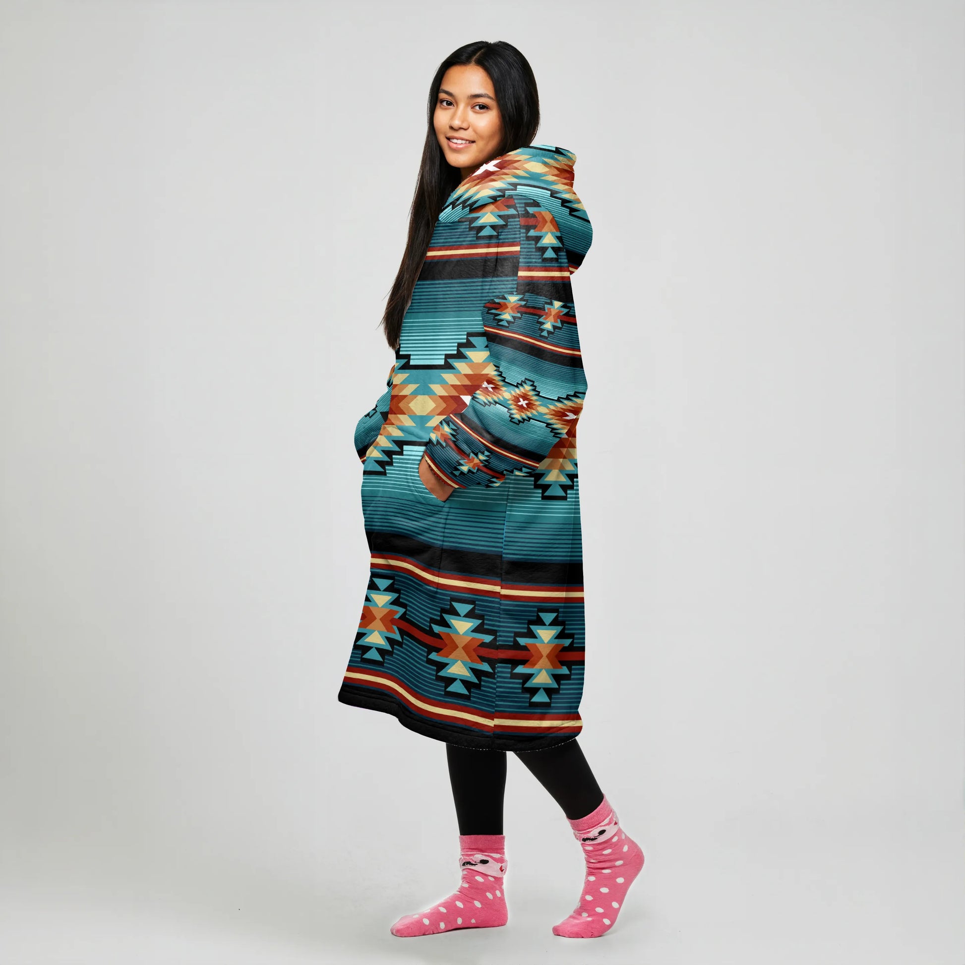 A woman front-facing in an oversized teal and coral striped tribal blanket hoodie, hands tucked into the pocket, smiling at the camera.