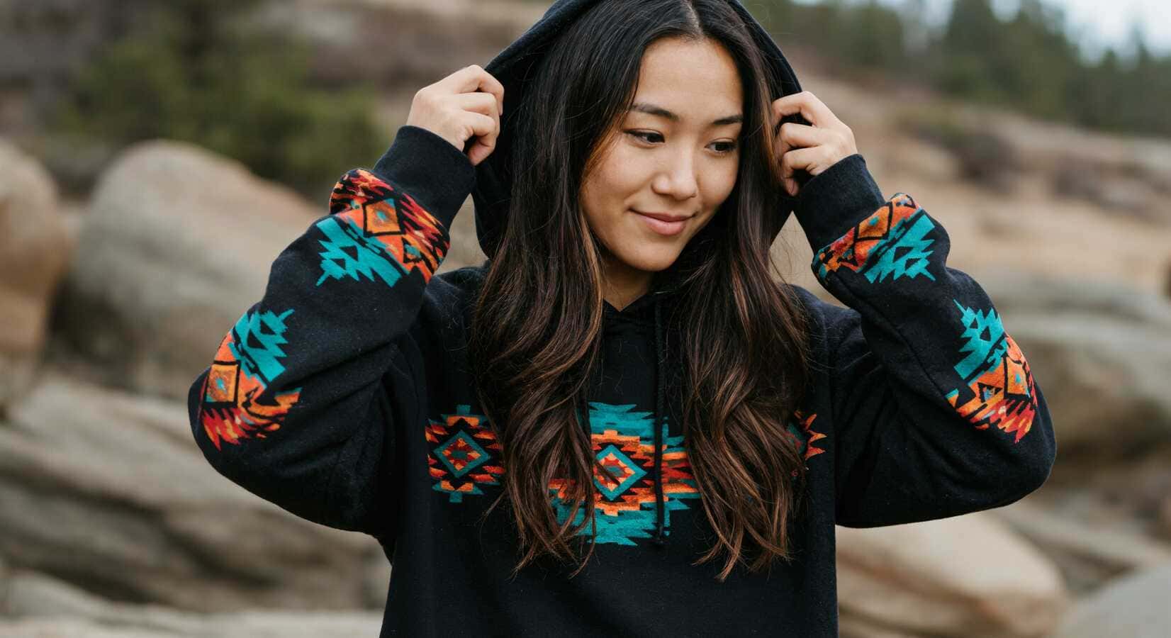 A young woman standing among rocks outdoors, wearing a black hoodie emblazoned with a colorful Southwestern-inspired geometric pattern as she gently pulls the hood over her head.1621243260e1af0c20-0