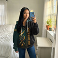 oman taking a mirror selfie wearing a black zip-up jacket featuring a vertical dreamcatcher design entwined with a tribal warbonnet motif, set against a dark green background on one half and solid black on the other.