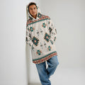 
Front view of a unisex cream wearable blanket hoodie featuring green geometric Native-American motifs, hand in pocket, styled with relaxed denim against white wall