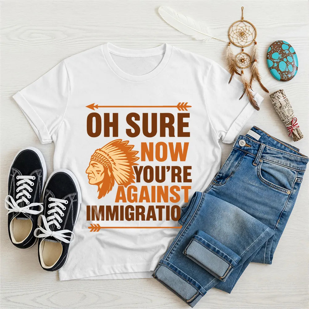 White t‑shirt featuring brown chief headdress graphic and text “Oh Sure Now Youre Against Immigration.”