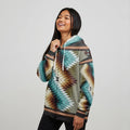 Female model wearing a teal Native-American tribal print zip-up jacket with bold geometric motifs and kangaroo pocket, posing against a seamless white background
