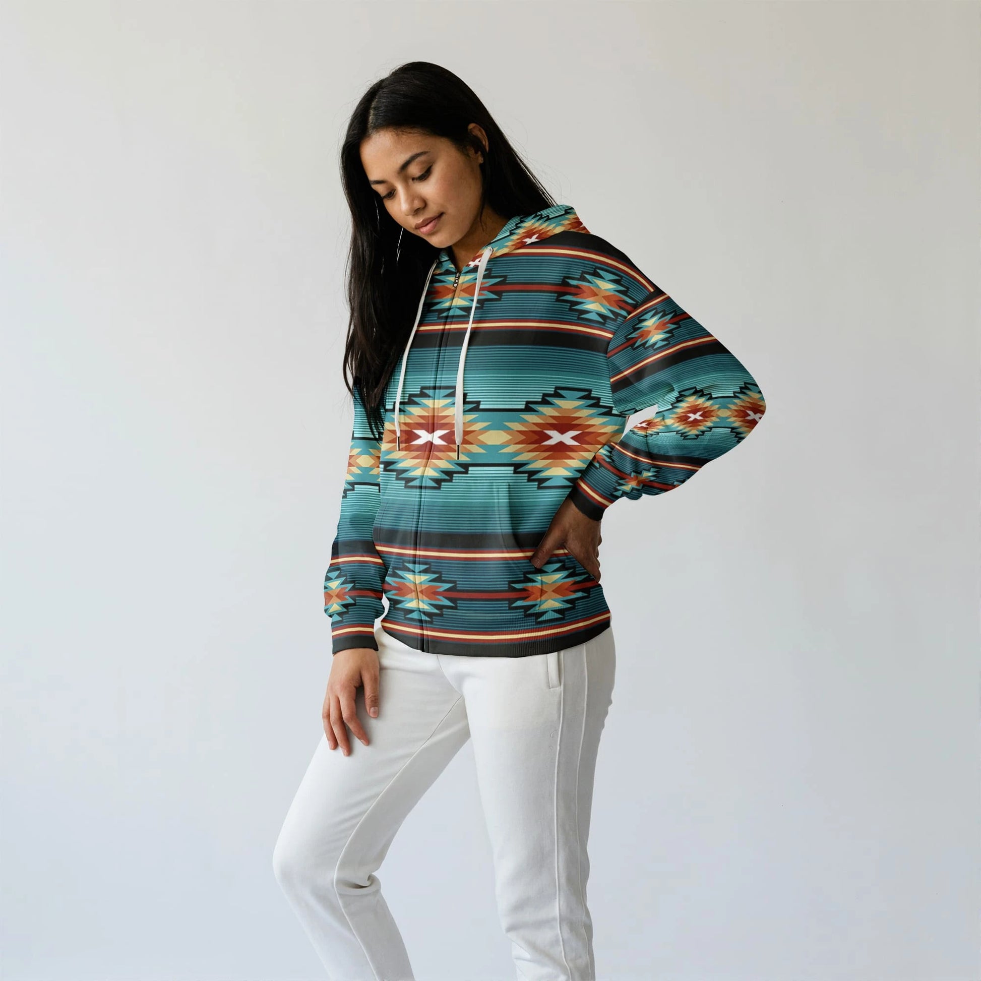 Side-angle of a female model wearing a teal Native-American–inspired pullover hoodie with kangaroo pocket, head tilted down, hands in pocket, on a plain white backdrop