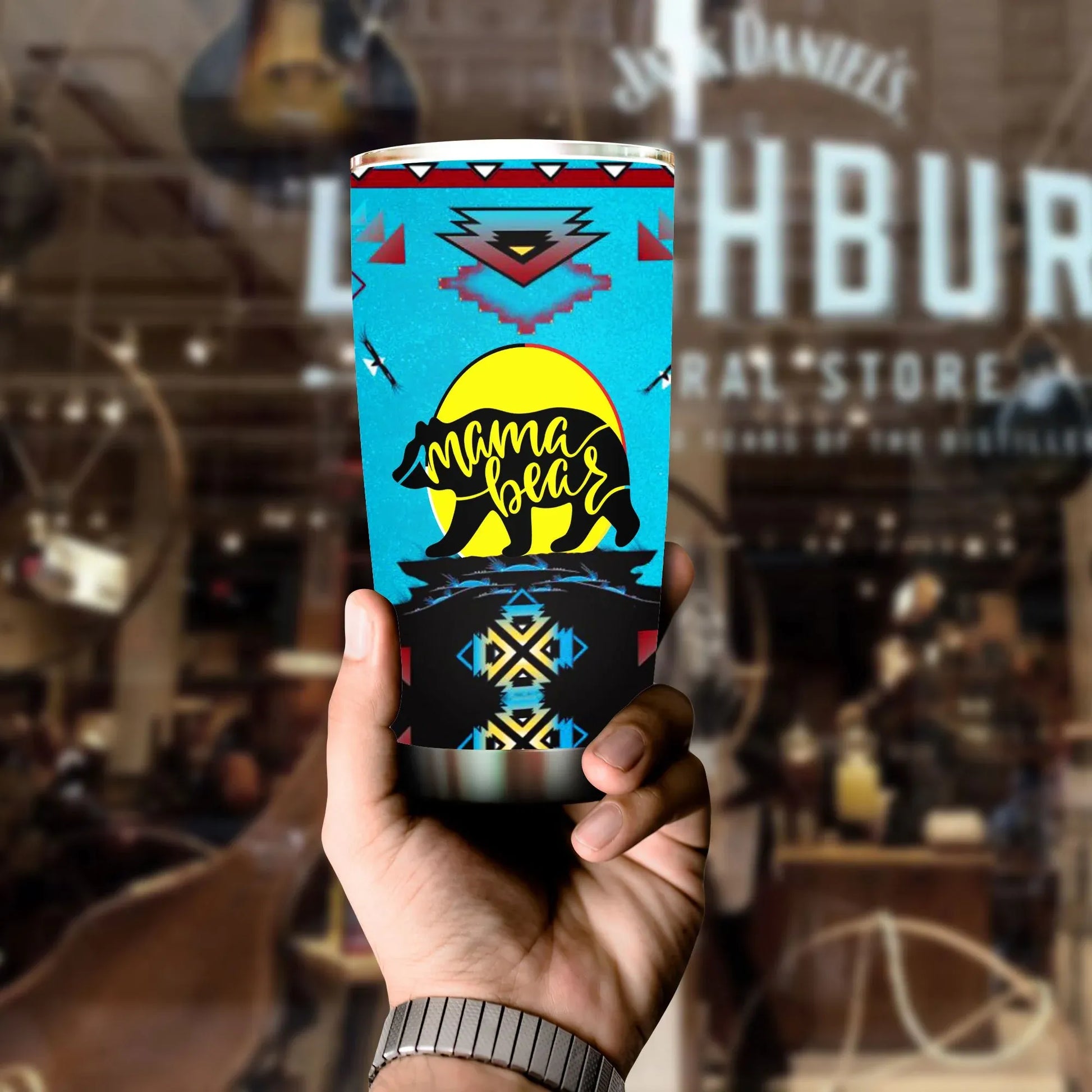 Hand holding Mama Bear tumbler with bright blue and black Native American style pattern, blurred bar background.
