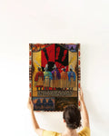 Hands hanging a framed Plains‑inspired canvas print on a white wall, featuring five tribal dancers in regalia before a red‑and‑black handprint and tipi border.