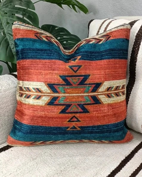 Rust and teal Southwestern Aztec pattern throw pillow sitting on a cream sofa next to a leafy houseplant.