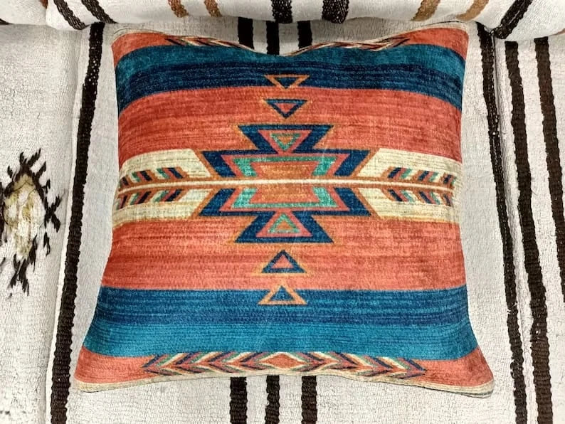 Top-down view of a rust & teal Southwestern Aztec throw pillow resting on a textured blanket.