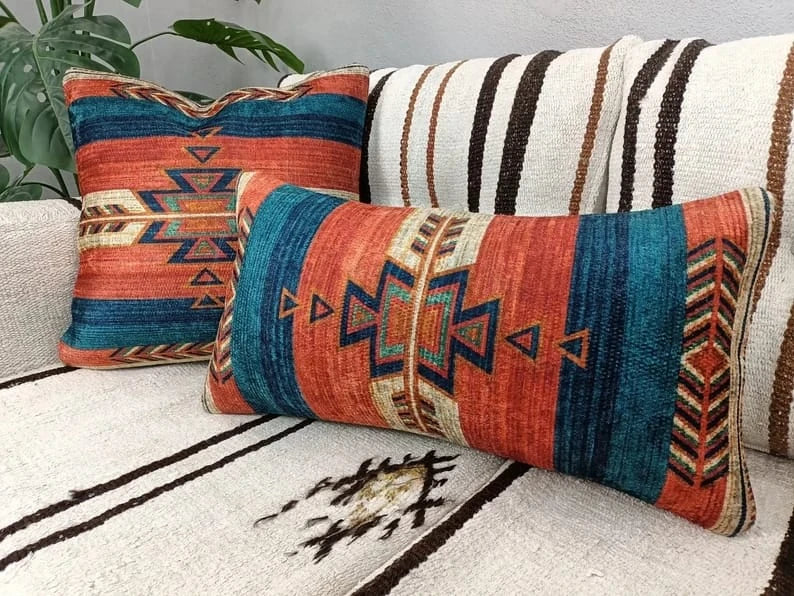 Rust and teal Southwestern Aztec pattern throw pillow sitting on a cream sofa next to a leafy houseplant.