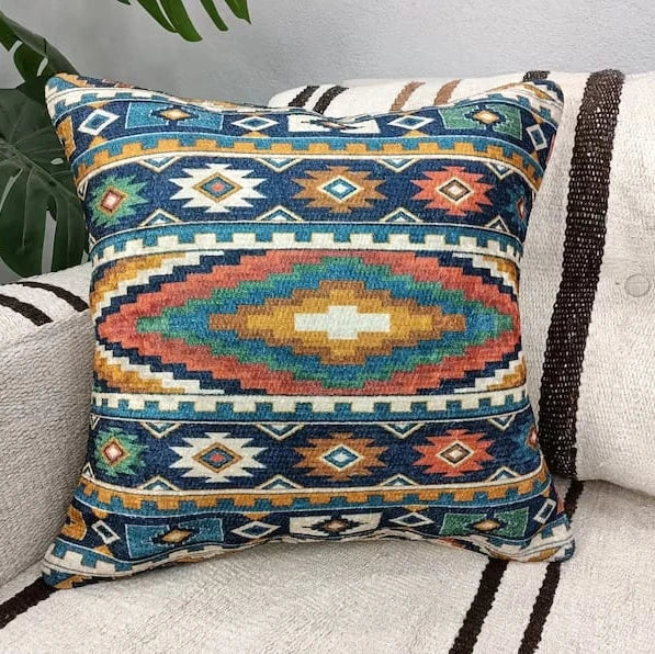 Square teal, rust and gold Aztec pattern throw pillow resting on a cream sofa next to a large leafy plant.