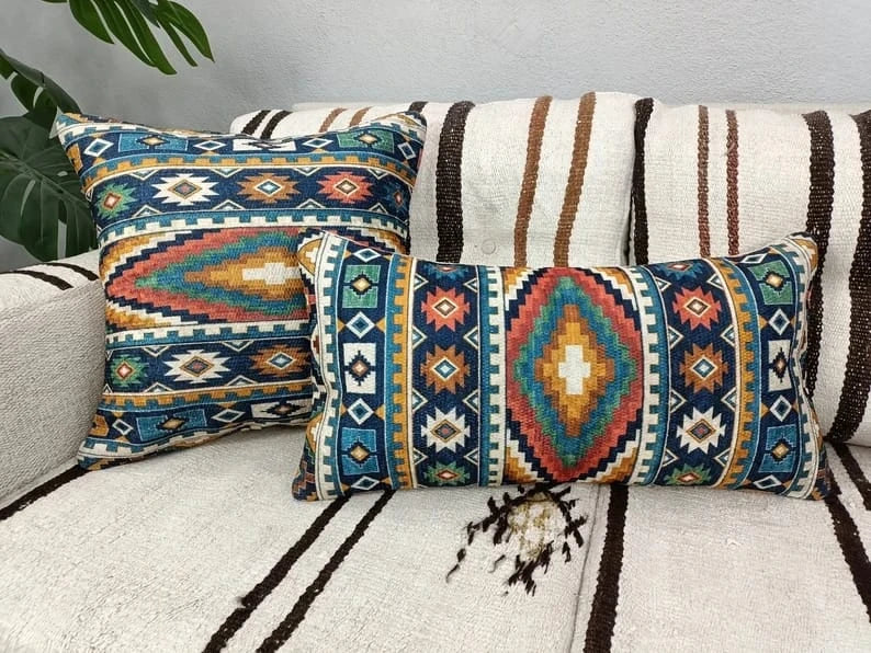 Square teal, rust and gold Aztec pattern throw pillow resting on a cream sofa next to a large leafy plant.