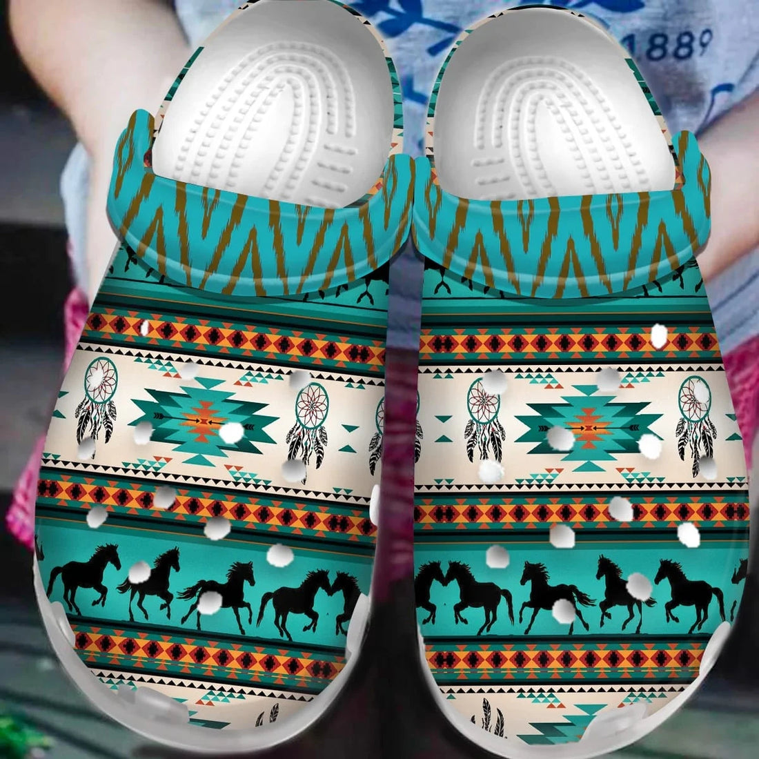 Pair of clog mules with turquoise, rust and cream Southwestern dreamcatcher and horse print held outdoors by a person’s hands.