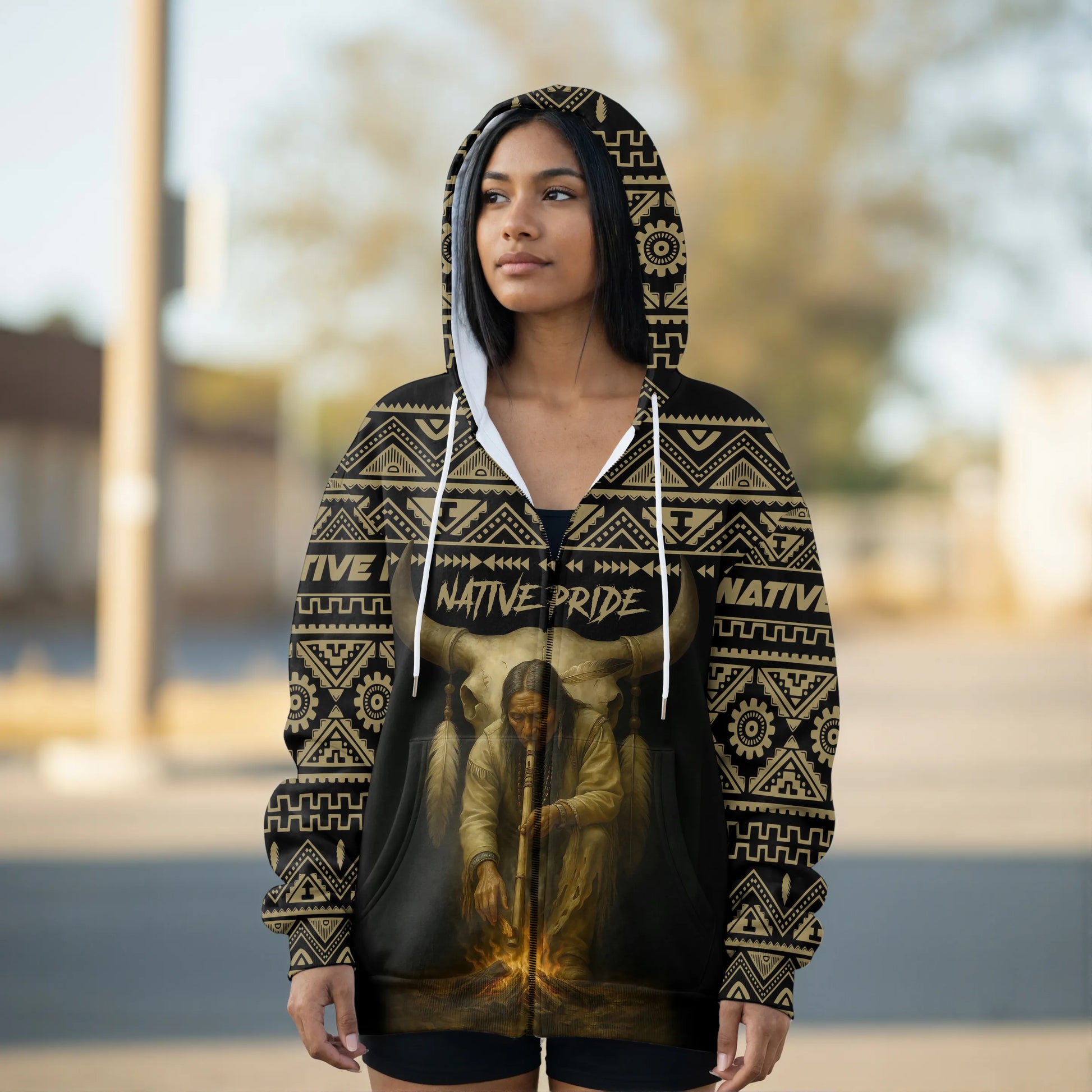 Front of a black zip-up hoodie with gold geometric tribal designs on the hood and shoulders, showing the same Native elder flute-player and buffalo skull artwork across the chest, a full-length zipper, and kangaroo pockets, embodying a vintage Indigenous pride aesthetic.