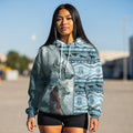 Female model standing in sunlight wearing a sky-blue hoodie split diagonally between a lifelike 3D wolf portrait and a Native warrior scene, the dreamcatcher and running-wolf pattern covering the opposite side and extending onto the sleeves and hood.